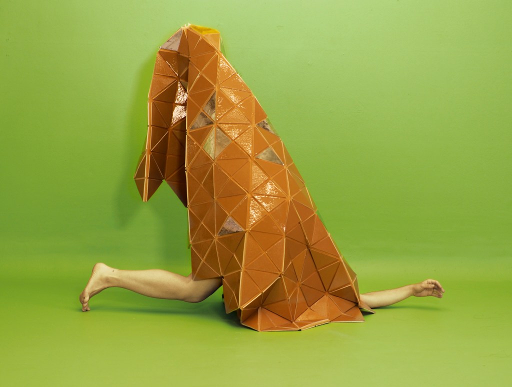 A rust colored solid patchwork quilt-like sculpture with the dancer's lower leg and one forearm protruding, on a green background.