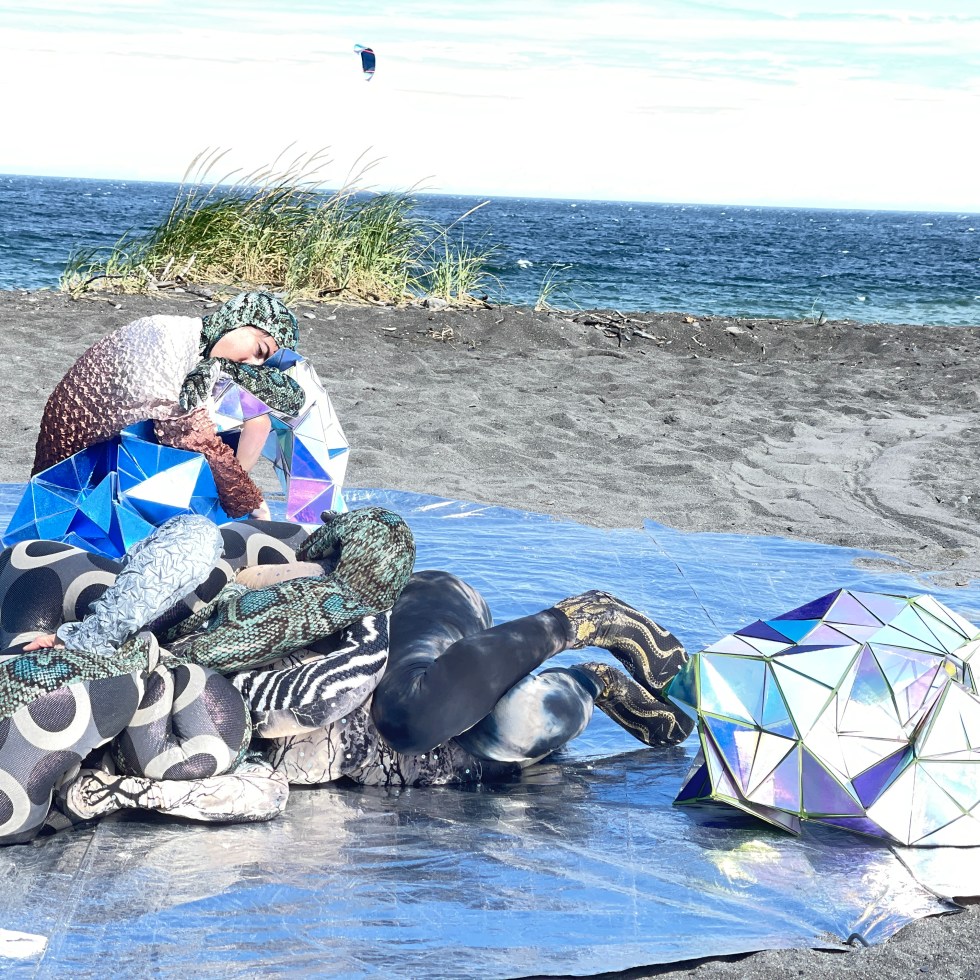 Two dancers tangled with soft snake-like sculptures and geodesic quilt sculptures, on a grey sand beach.