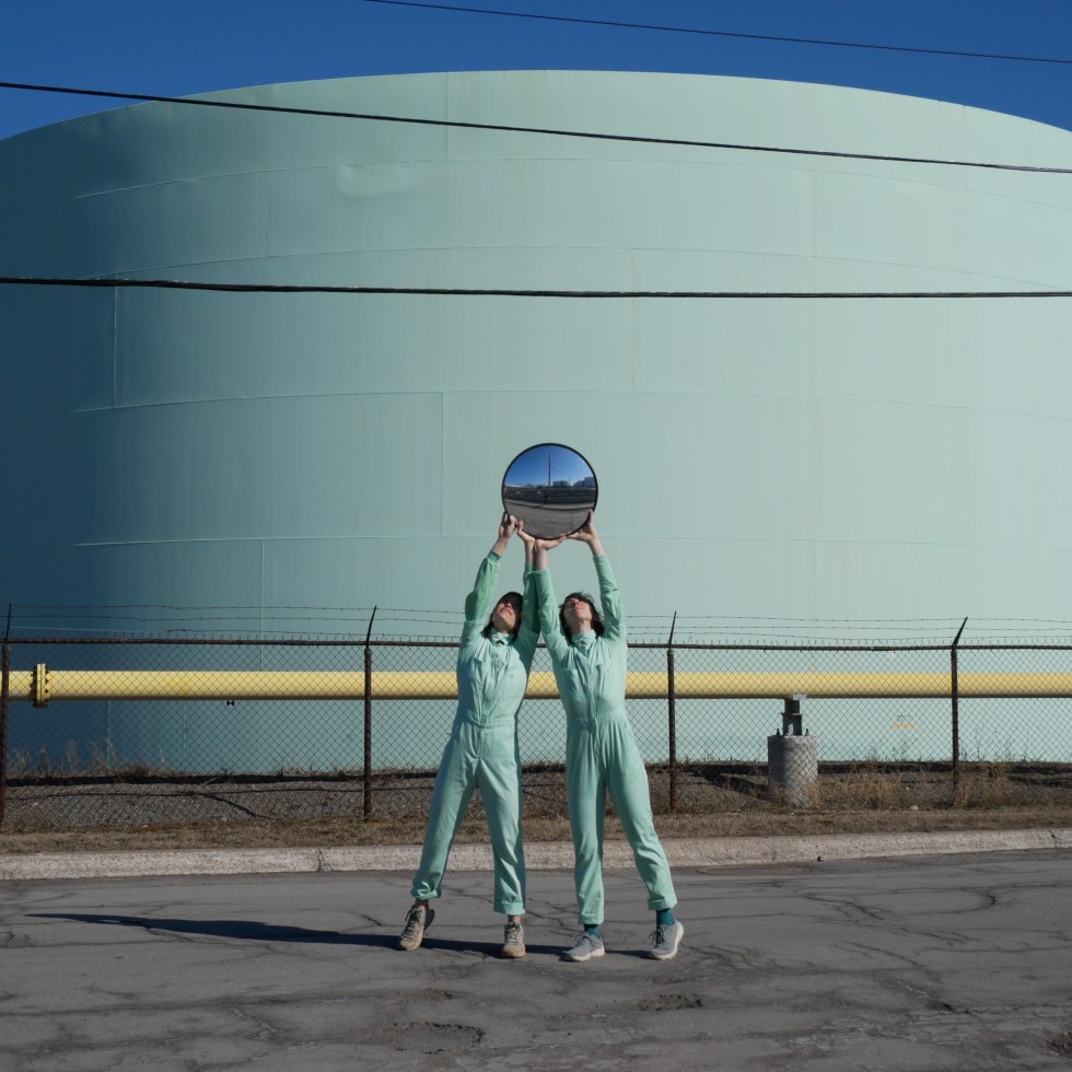 Lucy and Isabella hold a convex security mirror together high above their heads, the Suncor refinery behind them.